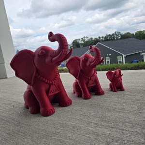 May include: Three matte red elephant figurines of varying sizes, each featuring a beaded necklace with a triangular pendant displaying the Greek letters Delta Sigma Theta. The elephants are depicted in a seated position with trunks raised upward.