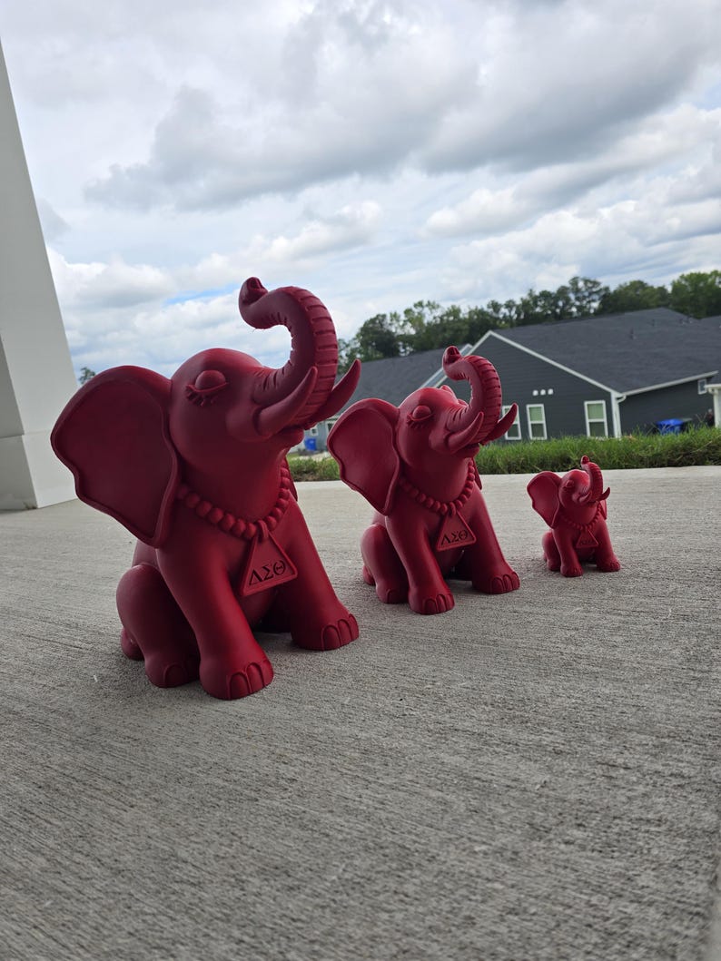 May include: Three matte red elephant figurines of varying sizes, each featuring a beaded necklace with a triangular pendant displaying the Greek letters Delta Sigma Theta. The elephants are depicted in a seated position with trunks raised upward.