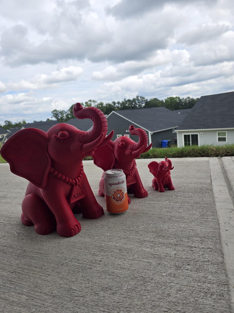 May include: Three matte red elephant figurines of varying sizes arranged in a row. Each elephant features a necklace with a triangular pendant displaying the Greek letters Delta Sigma Theta. A Spindrift Blood Orange sparkling water can is placed next to the largest elephant for scale.