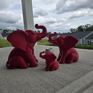 May include: Three matte red elephant figurines of varying sizes arranged in a group. The two larger elephants have trunks raised upward and closed eyes, while the smallest elephant sits in the center. A silver soda can is placed behind the smallest.