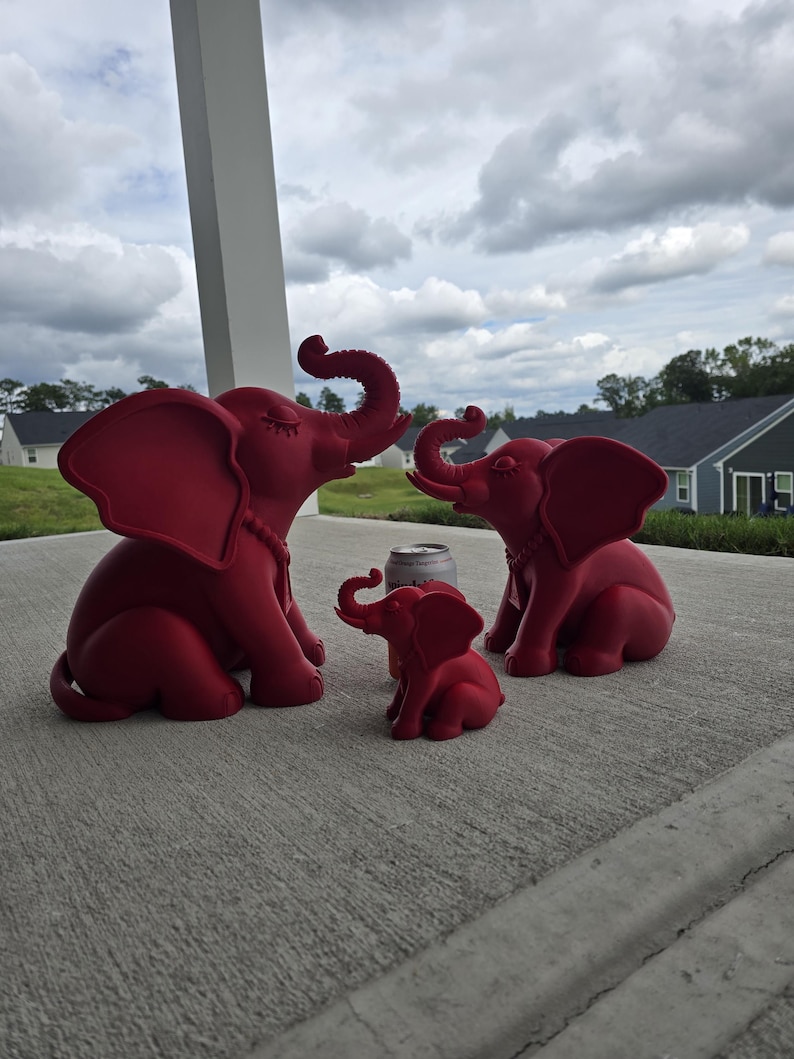 May include: Three matte red elephant figurines of varying sizes arranged in a group. The two larger elephants have trunks raised upward and closed eyes, while the smallest elephant sits in the center. A silver soda can is placed behind the smallest.