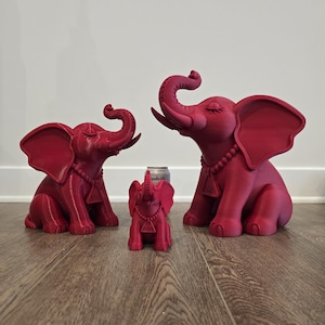 May include: Three matte dark pink elephant figurines of varying sizes, each featuring a beaded necklace with a triangular pendant. The elephants are depicted in a seated position with trunks raised upward and closed eyes, arranged in a row.