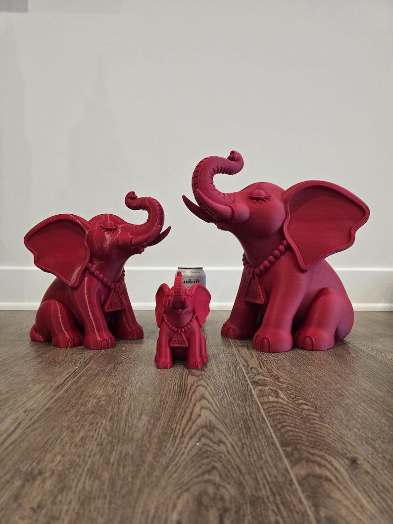 May include: Three matte dark pink elephant figurines of varying sizes, each featuring a beaded necklace with a triangular pendant. The elephants are depicted in a seated position with trunks raised upward and closed eyes, arranged in a row.