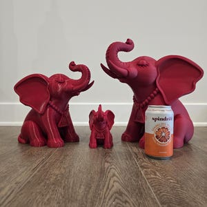 May include: Three matte magenta elephant figurines in varying sizes, each featuring a beaded necklace with a triangular pendant bearing the Greek letters Delta and Theta. A 12-ounce Spindrift Blood Orange sparkling water can is placed for scale.