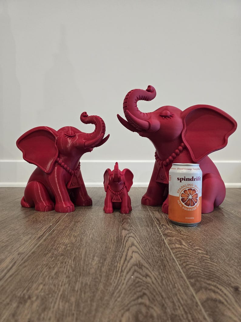 May include: Three matte magenta elephant figurines in varying sizes, each featuring a beaded necklace with a triangular pendant bearing the Greek letters Delta and Theta. A 12-ounce Spindrift Blood Orange sparkling water can is placed for scale.