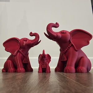 May include: Three matte crimson elephant figurines of varying sizes, each featuring a beaded necklace with a triangular pendant displaying the Greek letters Delta and Theta. The elephants are depicted in a seated position with trunks raised.