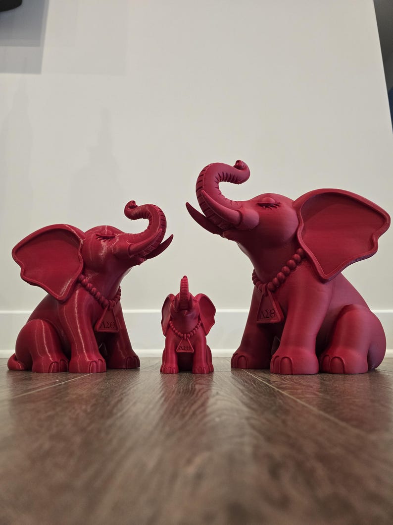 May include: Three matte crimson elephant figurines of varying sizes, each featuring a beaded necklace with a triangular pendant displaying the Greek letters Delta and Theta. The elephants are depicted in a seated position with trunks raised.