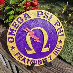 May include: A circular sign in purple and gold, featuring the words "OMEGA PSI PHI FRATERNITY INC". The centre has a gold Omega symbol with a red lightning bolt and the year "1911". The sign is displayed on a wooden surface.
