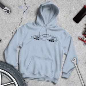 May include: A pale blue hoodie with a black outline of a car. The image includes car parts such as a tyre, spanners, and a shock absorber. The background is a concrete surface.