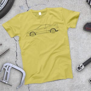 Puede incluir: Una camiseta amarilla con el contorno negro de un coche en la parte delantera.