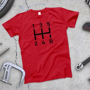 May include: Red t-shirt with a black graphic of a car's gear shift pattern. The numbers 1, 2, 3, 4, 5, and R are displayed on the gear shift.