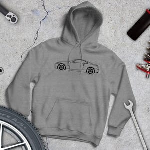 May include: A gray hoodie featuring a black line drawing of a sports car. The hoodie is surrounded by automotive tools and a tire, suggesting a car enthusiast theme. The background is a concrete surface.