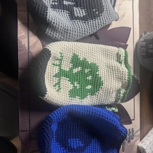 Könnte beinhalten: Drei gehäkelte Mützen. Die obere Mütze ist grau mit einem dunkelgrauen Design. Die mittlere Mütze ist weiß mit einem grünen Design. Die untere Mütze ist blau. Alle Mützen sind handgefertigt.