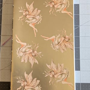 May include: A gold-colored notebook featuring illustrations of fairies with flowing hair and large wings. The fairies are depicted in various poses, with soft peach and cream tones. The notebook has a white spine and is lying on a gray cutting mat.
