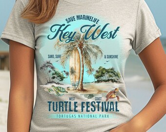 May include: A light gray t-shirt featuring a beach scene with palm trees, a surfboard, and turtles. The text on the shirt reads "Save Marine Life Key West Turtle Festival" with "Sand, Surf & Sunshine" below. The shirt is worn by a person.