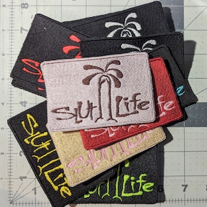 Op de afbeelding: Een stapel van acht geborduurde patches met de tekst "Slut Life" in verschillende kleuren en lettertypen. De patches hebben een palmboomontwerp en zijn in een verspringende volgorde gerangschikt.