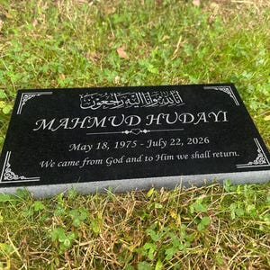 May include: A black granite memorial plaque with white engraved text. The inscription includes the name "MAHMUD HUDAYI", dates, and a religious quote. Decorative corner designs frame the text. The plaque rests on grass.