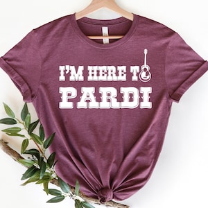Puede incluir: Camiseta color burdeos con texto blanco que dice "I'm here to PARDi" con un gráfico de guitarra.