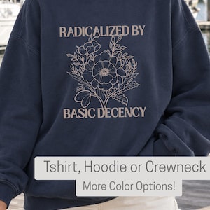 Puede incluir: Una sudadera de cuello redondo en color azul marino oscuro con una ilustración floral de líneas en color rosa claro en el centro. El texto RADICALIZED BY está impreso sobre las flores y el texto BASIC DECENCY debajo del diseño floral.
