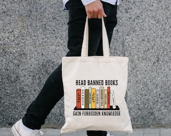 Puede incluir: Un bolso tote de color crema con el texto "READ BANNED BOOKS GAIN FORBIDDEN KNOWLEDGE" sobre un gráfico de libros coloridos. El bolso lo lleva una persona que lleva una camisa a rayas, pantalones negros y zapatillas blancas.