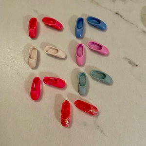 May include: A collection of miniature ballet slippers in various colors, including red, pink, blue, and a pale beige. Some slippers have glitter accents. The shoes are arranged on a light-colored surface.