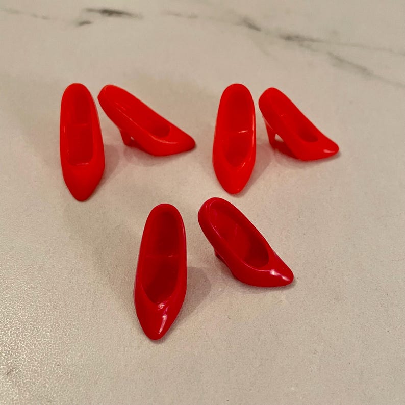 May include: Six red plastic high-heeled shoes for a doll. The shoes are all the same style and are arranged in a scattered pattern on a white surface.