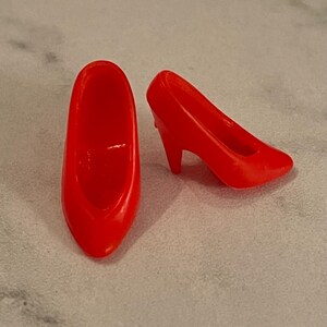 May include: A pair of bright red high-heeled shoes for a doll. The shoes are made of plastic and have a simple design.