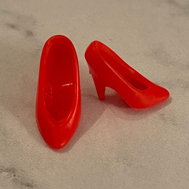 May include: A pair of bright red high-heeled shoes for a doll. The shoes are made of plastic and have a simple design.