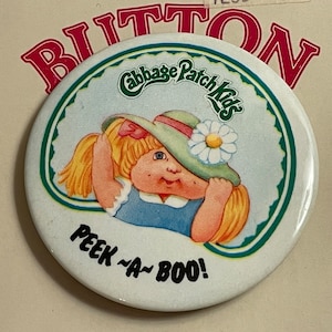 May include: A white round button with a green border. The button features a Cabbage Patch Kids doll with blonde hair wearing a green hat with a white flower. The text "Cabbage Patch Kids" is written in green letters on a white background. The text "Peek-a-Boo!" is written in black letters on a white background.