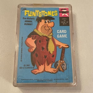 May include: A blue and yellow card game box featuring Fred Flintstone from the cartoon "The Flintstones". The box features the text "The Flintstones Pre-Historic Animal Rummy Games to Grow Up With Card Game".
