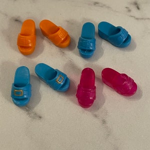 May include: Six pairs of plastic doll shoes in orange, blue and pink. The shoes are all slip-on sandals with a buckle detail.