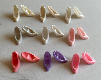 May include: A collection of 18 miniature plastic doll shoes in various colors, including white, pink, and purple. The shoes are arranged in three rows on a white surface.