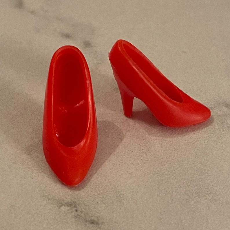 May include: A pair of red high-heeled shoes for a doll. The shoes are made of plastic and have a simple design.