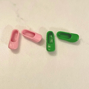 May include: Four miniature clogs in pink and green. Two pink clogs are positioned on the left, while two green clogs are on the right. The clogs are made of a smooth material and are designed to resemble traditional wooden shoes.