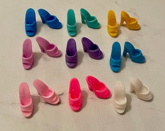 May include: A collection of 12 pairs of colorful plastic doll shoes. The shoes are all different colors, including pink, blue, purple, yellow, and white. The shoes are all made of plastic and have a simple design.