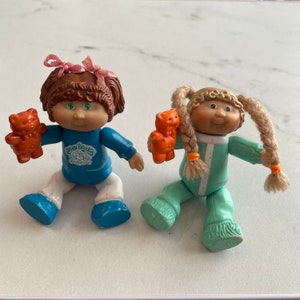 以下が含まれることがあります： パジャマを着て、小さなオレンジ色のぬいぐるみを持ったキャベッジパッチキッズ人形が2体。左の人形は、キャベッジパッチキッズのロゴが入った青いシャツと白いズボンを着用しています。右の人形は、緑と白のストライプのパジャマを着ています。