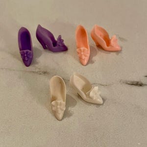 May include: Six pairs of doll shoes in pink, purple and white with a flower detail. The shoes are all different styles and sizes.