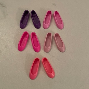 May include: Set of ten pairs of doll shoes in various shades of pink and purple. The shoes are made of plastic and have a simple, classic design.