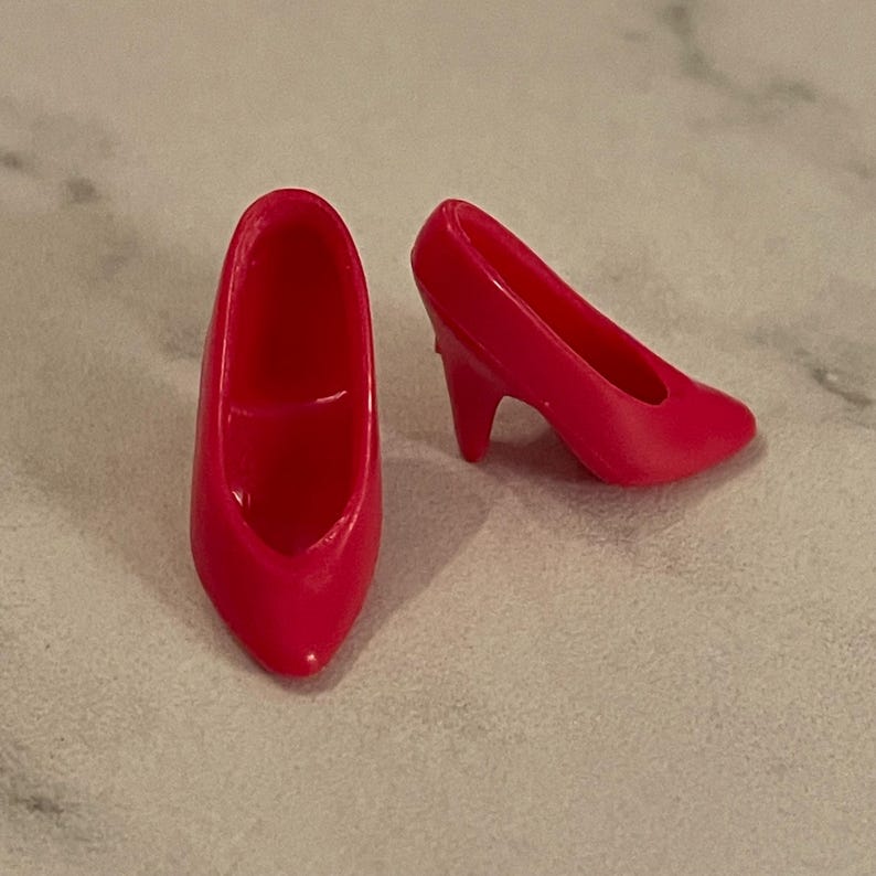 May include: A pair of bright pink high-heeled shoes for a doll. The shoes are made of plastic and have a pointed toe.