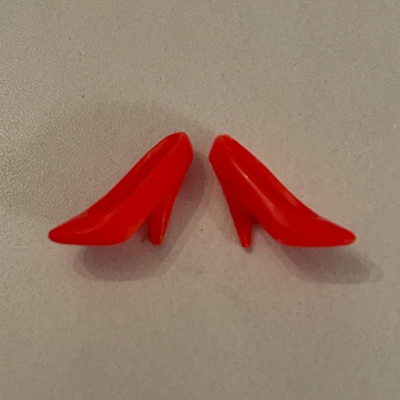 May include: A pair of bright red high-heeled shoes made of plastic. The shoes are pointed-toe and have a simple design.