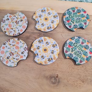 May include: Set of six car coasters with various designs. Two coasters feature a floral and bee pattern, two have a sunflower and bee design, and two display a cactus and the text "Grow Vibes". Each coaster has a black border.