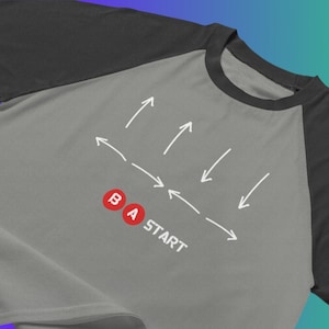 Może przedstawiać: Szara i czarna koszulka bejsbolowa z białym wzorem strzałek wskazujących na słowa "BA START" i dwa czerwone koła z literami "B" i "A" w środku.