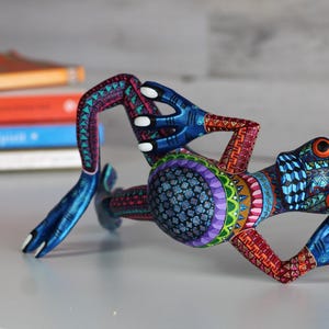May include: A brightly colored wooden frog figurine with blue, green, red, purple, and yellow paint. The frog is lying on its back with its legs outstretched.