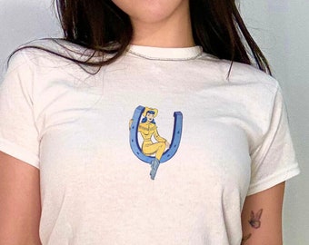 Może przedstawiać: Biały t-shirt z grafiką kobiety siedzącej na żółtej podkowie. Kobieta ma na sobie żółty strój. T-shirt jest połączony z białymi spodniami. Srebrna torebka jest trzymana w dłoni.