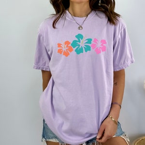 May include: A lavender t-shirt with a graphic of three hibiscus flowers in pink, orange, and teal.
