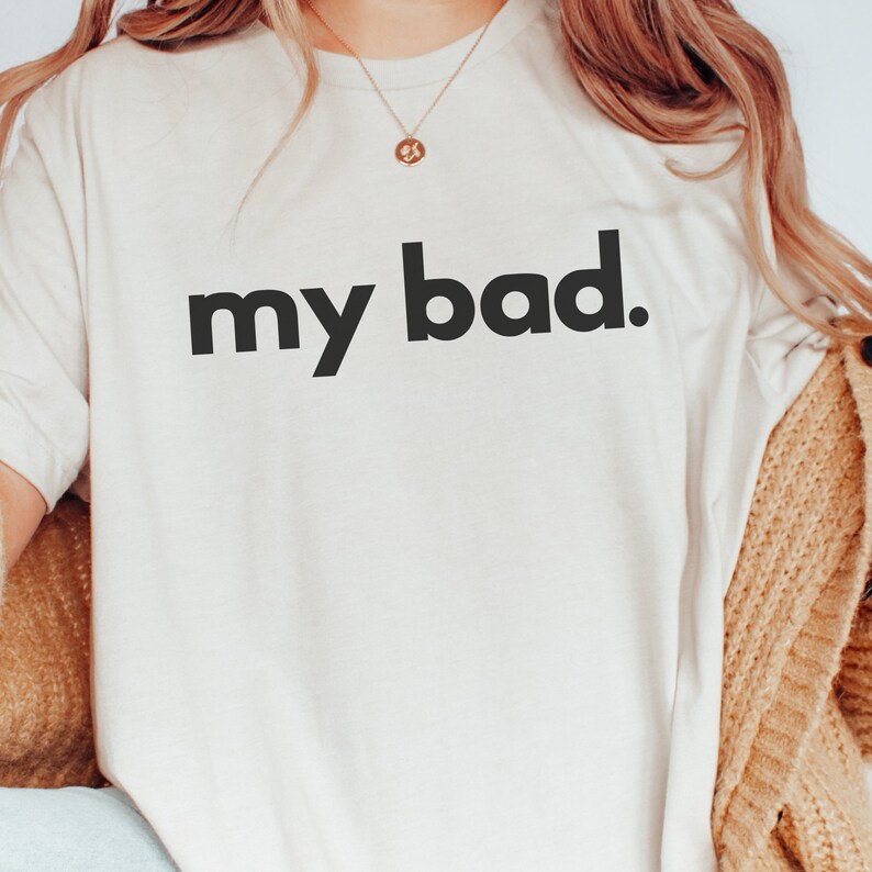 May include: Cream-colored t-shirt with the phrase "my bad." printed in bold, black letters. The shirt is a simple crew neck style, and the text is centered on the front.