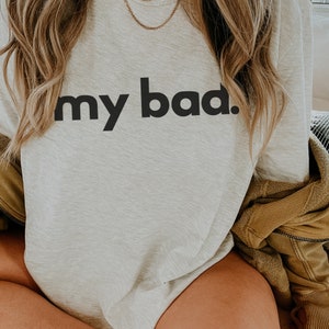 May include: A light gray sweatshirt with the phrase "my bad." printed in bold, black letters. The sweatshirt is a casual style, suitable for everyday wear. The image shows the top of the sweatshirt and part of the person wearing it.