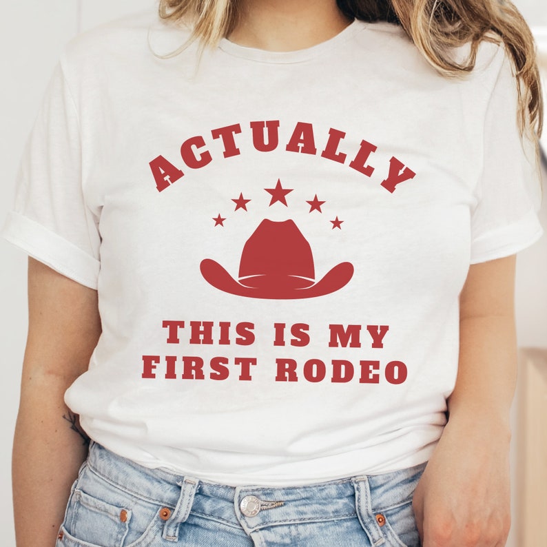 K&ouml;nnte beinhalten: Wei&szlig;es T-Shirt mit rotem Text, der "Actually This Is My First Rodeo" lautet, mit einem roten Cowboyhut und Sternen &uuml;ber dem Text.