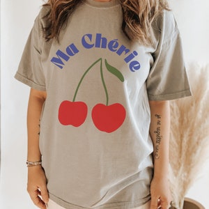 May include: A gray t-shirt with the text "Ma Ch&eacute;rie" in blue letters and a graphic of two red cherries. The text "je ne regrette rien" is written in black on the sleeve.
