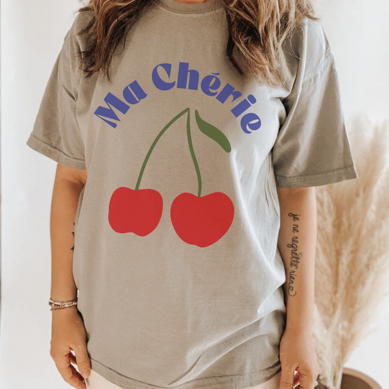 May include: A gray t-shirt with the text "Ma Ch&eacute;rie" in blue letters and a graphic of two red cherries. The text "je ne regrette rien" is written in black on the sleeve.
